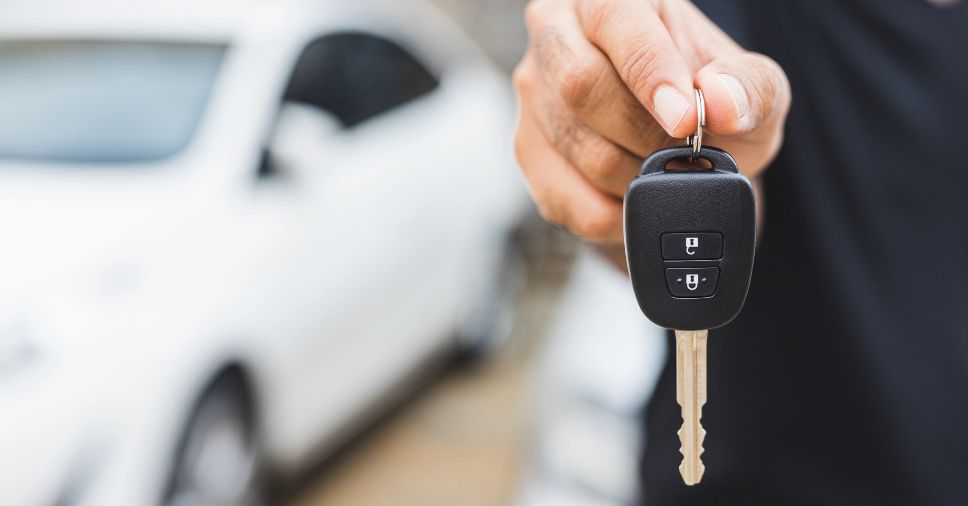 Mão segura chave de carro com veículo ao fundo, ilustrando a consulta de histórico veicular.